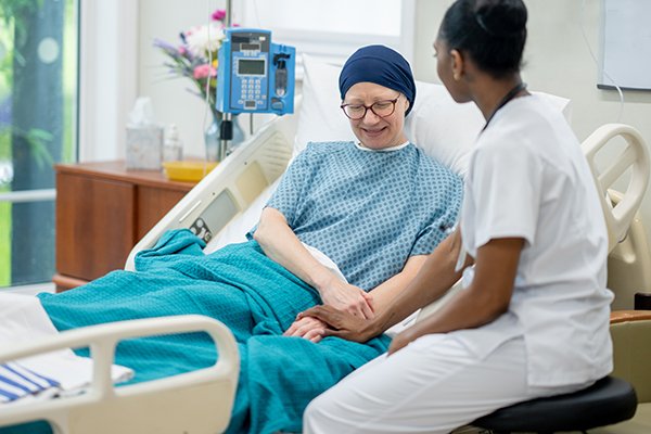 A nurse sits at the bedside of an older female oncology patient wearing a head scarf
