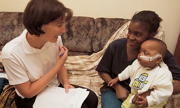 A health professional on a home visit talks to a mother holding a toddler who has a nasogastric tube in place