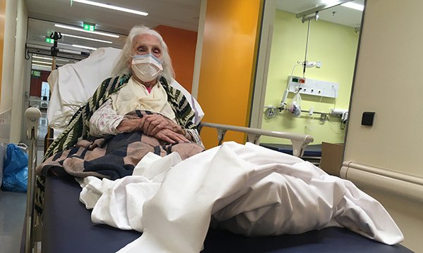 Older woman lies in bed in corridor of emergency department