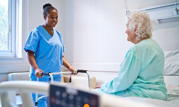 An older woman in hospital sits on her bed and a nurse has brought a walking frame to encourage her to move, as this will help manage deconditioning syndrome