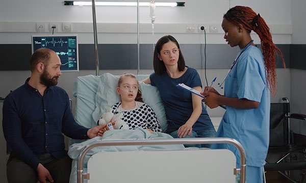 A nurse talks to a mother sitting on the edge of her child’s hospital bed while the father, seated on the other side of the bed, holds his daughter’s hand