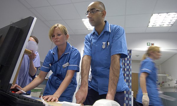 Two nurses look at a laptop screen together amid the bustle of a ward around them