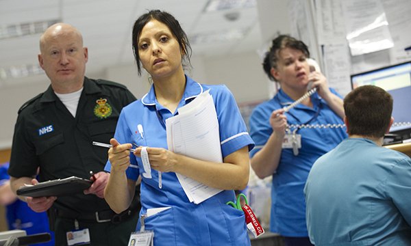 A busy emergency department with a paramedic looking serious, a nurse looking concerned while holding paperwork, while another nurse speaks to someone on the phone