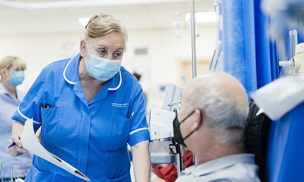 A nurse wearing a blue face masks talks to a patient who is sitting up and is also wearing a face mask