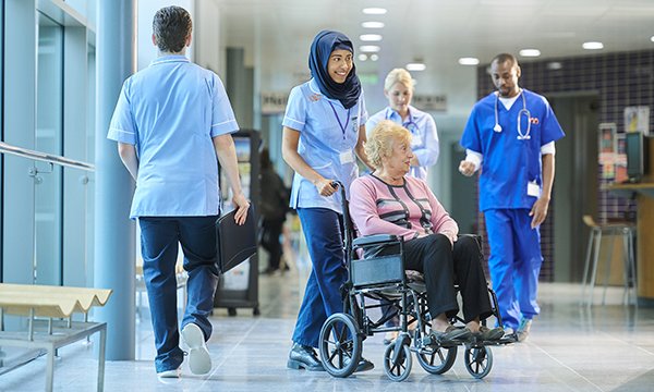 A nurse smiles and pushes an older woman in a wheelchair in a busy hospital setting