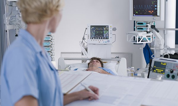 A nurse in ICU looks up from writing in notes to observe the patient. ICU can lack understanding of patients’ mental health care needs