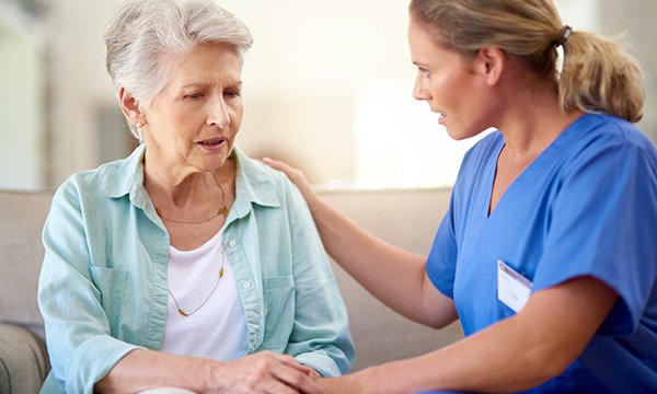 A nurse sits alongside an older woman patient who looks worried and puts a hand on her shoulder in a gesture of reassurance