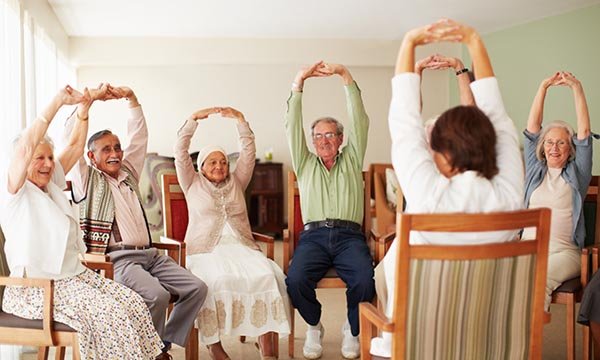 A group of older people do seated exercises with their hands above their heads following a seated instructor facing them