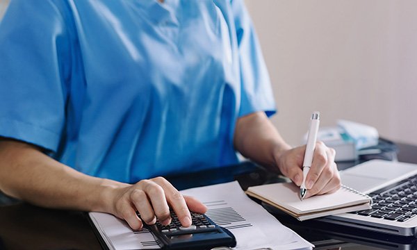 A nurse sitting at a table and making notes while using a calculator that is on top of a pile of documents, as if checking a payslip