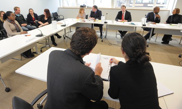 An NMC hearing with tables in a U shape, the panel members facing the camera and being addressed by two people sitting opposite them who are looking at a folder of documents