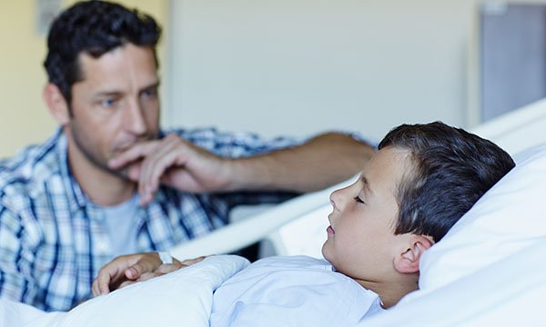 An anxious-looking father sits by his unconscious son’s hospital bedside watching him intently