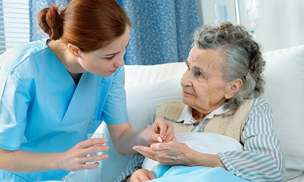 An older woman sitting up in bed holds out her hand to receive a pill from a nurse