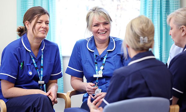 A group of nurses in uniform sit talking in a workplace setting, as if during a group clinical supervision session